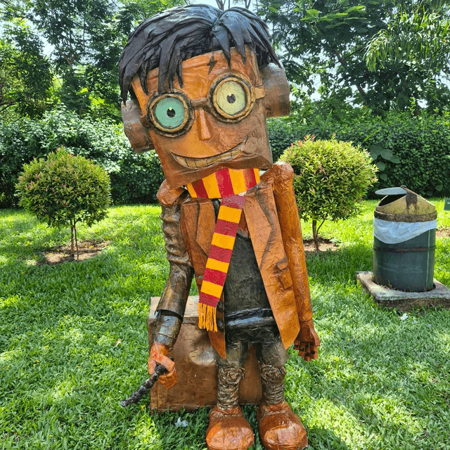 A carved figure with large glasses and a red-yellow scarf standing in a green campus garden.